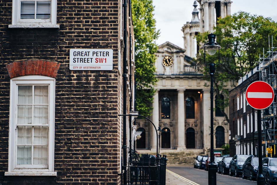 A close-up view of a brick building on Great Peter Street SW1 in Westminster, featuring a white-framed window with closed curtains, a street sign indicating 'GREAT PETER STREET SW1' from the City of Westminster, and a red and white 'no entry' traffic sign on the right. In the background, there is a historic church with large columns, a clock, and stone steps leading to its entrance. The scene is illuminated by natural daylight, with lush green trees partially visible, and parked cars lining the street. The overall atmosphere is calm and typical of a historic Westminster street scene, capturing the urban architecture and street signage.