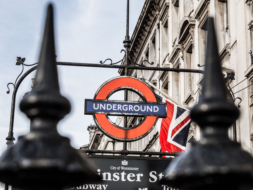 Close-up view of a London Underground station sign for Westminster, framed by black iron fencing in the foreground. The sign displays the classic red circle with a blue bar reading 'UNDERGROUND' and is mounted on a decorative metal arm attached to a historical building's facade, which features ornate stone detailing and large windows. A British flag is partially visible behind the sign, fluttering in the breeze. The scene is lit with natural daylight, and the overall atmosphere reflects an urban setting in Westminster, London, with a focus on public transit and city architecture. Westminster Carpet Cleaning does not appear in this image as it is a transportation-related scene.