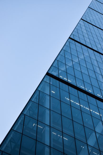 A close-up view of a modern office building's exterior featuring large glass windows with reflective blue-tinted surfaces, arranged in a grid pattern along the facade. The glass panels are clean and free of smudges, reflecting the clear sky above. The structure's sleek lines and sharp angles are highlighted by natural daylight, emphasizing the building's contemporary architectural design. The image captures part of the building's corner, with the windows extending vertically, indicating a multi-storey office space. Natural light illuminates the reflective surfaces, enhancing their shine and clarity. This visual showcases high-rise commercial building features typical of urban business districts, suitable for emphasizing surface cleaning and maintenance in professional environments. For thorough sanitisation and deep cleaning of similar glass facades and architectural surfaces, Westminster Carpet Cleaning offers expert commercial cleaning services, including surface cleaning, sanitisation, and taping for hygiene preservation in office settings.