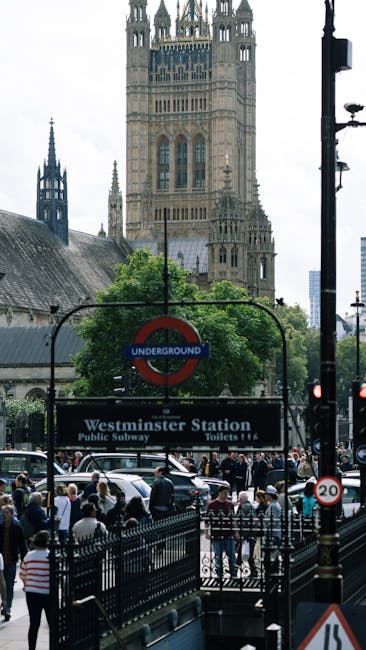 Close-up view of a London Underground station sign for Westminster, framed by black iron fencing in the foreground. The sign displays the classic red circle with a blue bar reading 'UNDERGROUND' and is mounted on a decorative metal arm attached to a historical building's facade, which features ornate stone detailing and large windows. A British flag is partially visible behind the sign, fluttering in the breeze. The scene is lit with natural daylight, and the overall atmosphere reflects an urban setting in Westminster, London, with a focus on public transit and city architecture. Westminster Carpet Cleaning does not appear in this image as it is a transportation-related scene.