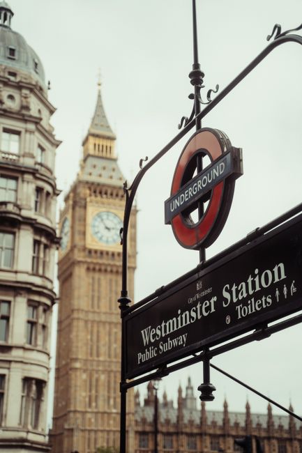 A close-up view of a black and red London Underground sign for Westminster Station, with the text 'Westminster Station' and symbols for toilets and public subway, mounted on an ornate black metal pole against the backdrop of historic buildings and the clock tower with Big Ben in the distance. The scene is outdoors with overcast skies, capturing the urban atmosphere of Westminster in London. Westminster Carpet Cleaning ensures thorough surface cleaning and sanitisation for residential and commercial properties in this area, maintaining hygiene and cleanliness standards.