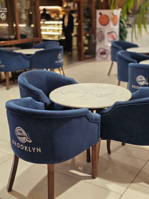 A set of four blue upholstered armchairs with wooden legs arranged around a round marble-topped table inside a cafe or shop setting. The chairs feature a logo with the word 'Brooklyn' and a stylized cityscape, indicating branding, and are situated on a light-colored tiled floor. In the background, partially visible shelves, display units, and some greenery add to the cozy atmosphere. The lighting appears natural and bright, highlighting the clean and well-maintained surfaces. The space is organized and tidy, reflecting professional deep cleaning, as provided by Westminster Carpet Cleaning, to ensure hygiene and freshness in commercial interiors.